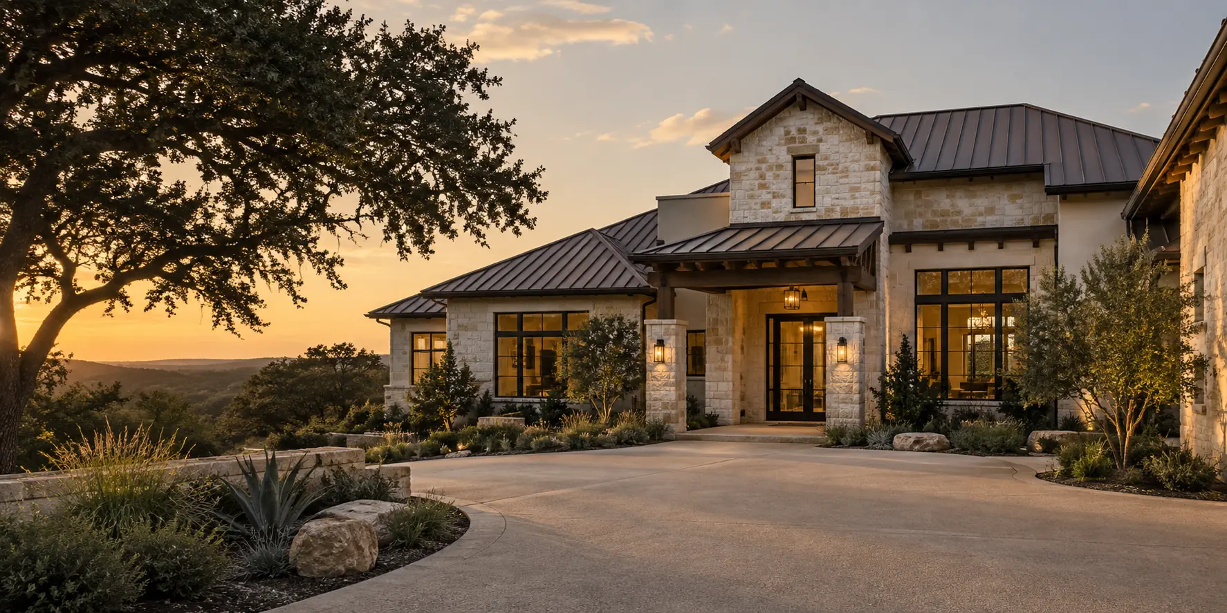 Limestone house with a dark standing-seam metal roof, covered entry and a live oak at sunset.